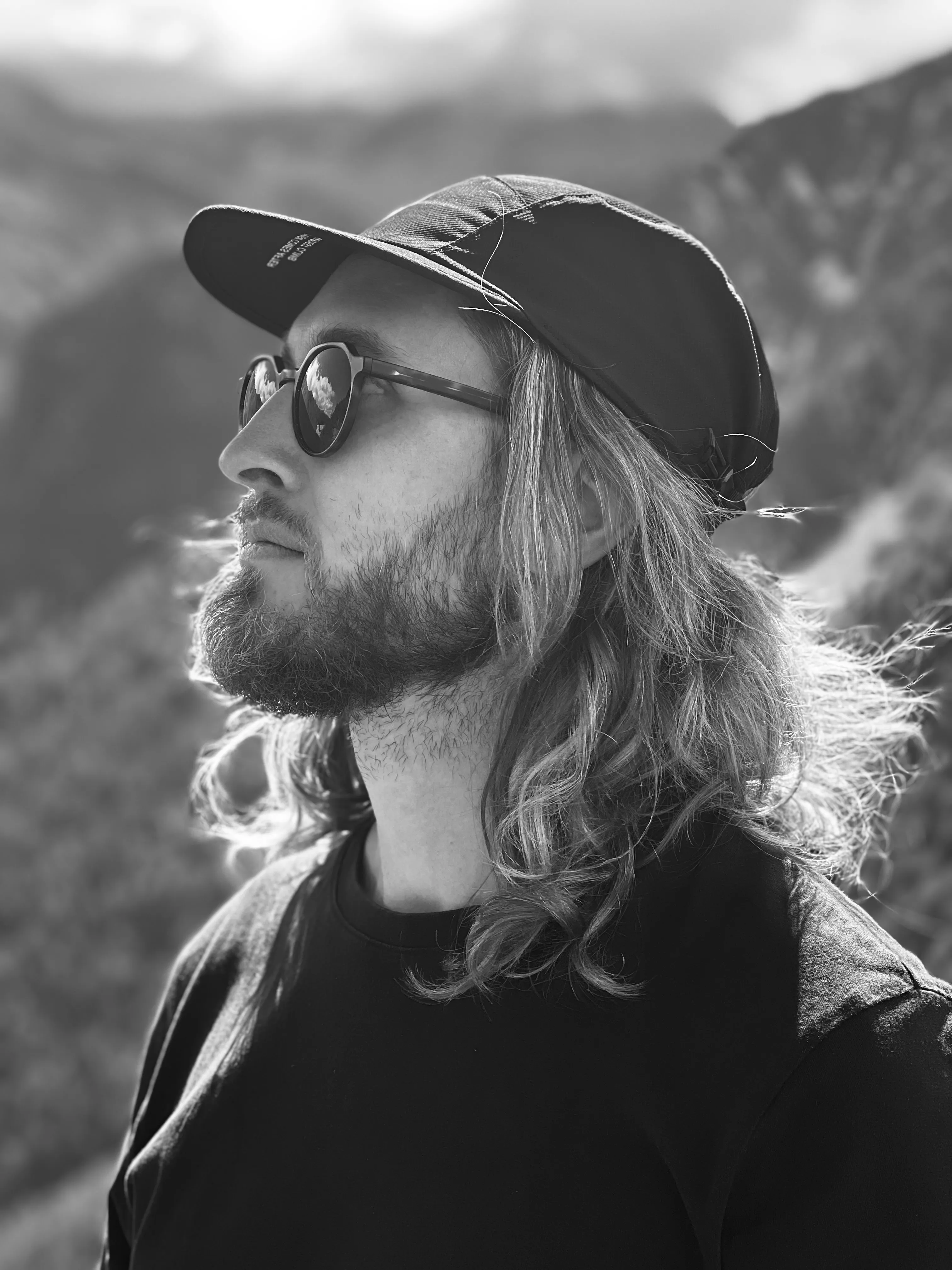 Alexander Müdespacher in profile, with long hair, a cap and sunglasses, in front of mountains.